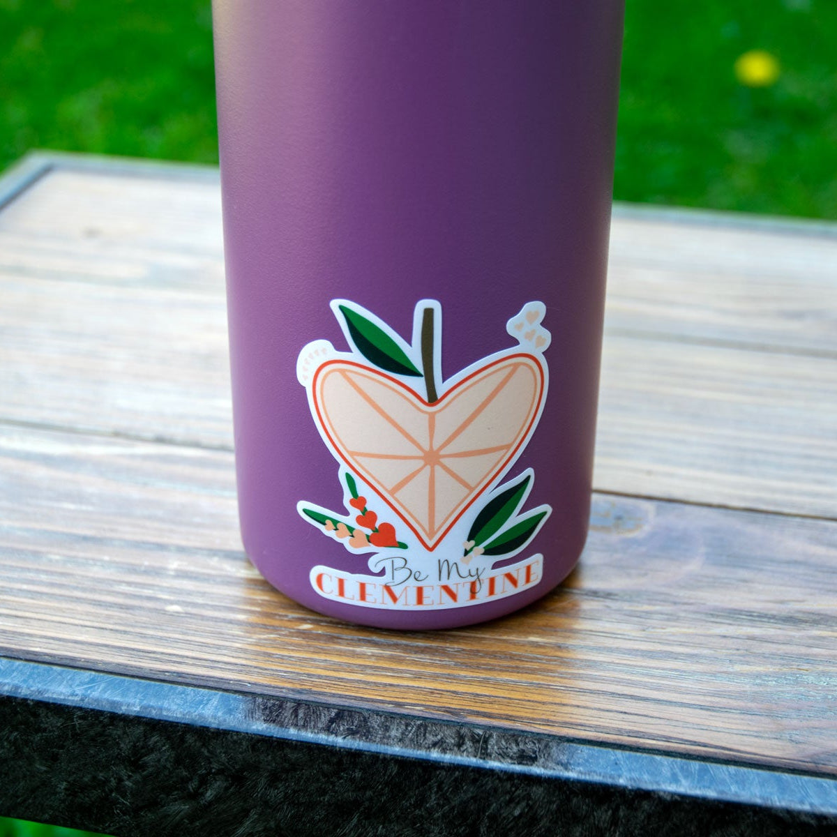 Heart sticker adorned qater bottle sitting on a wooden table on a daisy covered lawn.