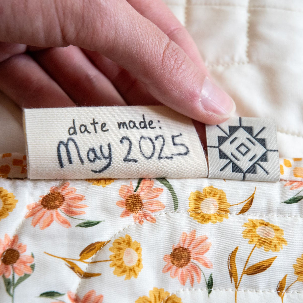 Hand holding a label with 'date made: May 2025' on floral-backed quilt.